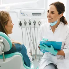 Uśmiechnięta dentystka z maską na szyi rozmawia z pacjentką. Czy do ortodonty potrzebne jest skierowanie? Dowiaduje się tego podczas wizyty.