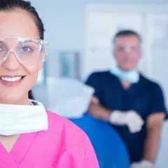 Uśmiechnięta asystentka dentysty w okularach ochronnych i różowym fartuchu, z maską na szyi. W tle widać lekarza.