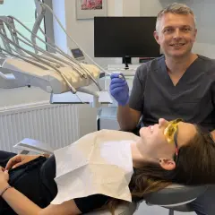 Dentysta w rękawiczce trzyma wiertło, pacjentka w okularach ochronnych leży na fotelu.
