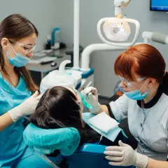 Dwie kobiety w okularach i maseczkach pracują przy pacjencie w gabinecie stomatologicznym. Jedna z nich, w niebieskim fartuchu, asystuje. To praktyczny element kursu asystentka stomatologiczna.