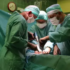 Trzech chirurgów w zielonych fartuchach i maseczkach wykonuje operację. Jeden z nich używa wiertarki chirurgicznej.