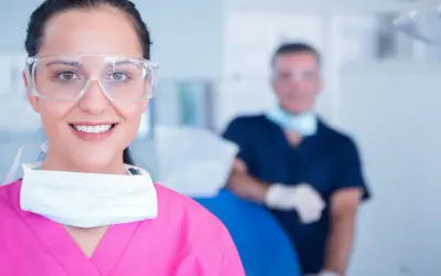 Uśmiechnięta asystentka dentysty w okularach ochronnych i różowym fartuchu, z maską na szyi. W tle widać lekarza.