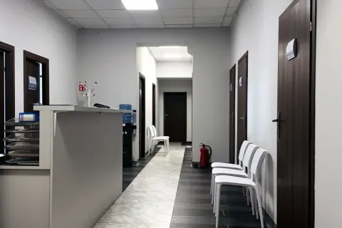 Korytarz przychodni z recepcją, drzwiami i białymi krzesłami.