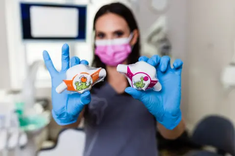 Dentystka w niebieskich rękawiczkach prezentuje dwa opakowania z zabawnymi dinozaurami, sugerujące zastosowanie w leczeniu, być może z użyciem podtlenku azotu.