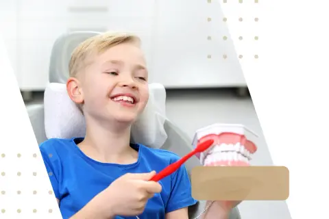 Uśmiechnięty chłopiec w gabinecie dentystycznym uczy się o higienie jamy ustnej, trzymając szczoteczkę i model zęba 26.