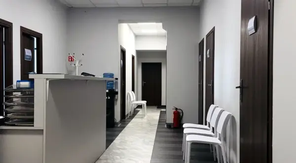 Korytarz przychodni z recepcją, drzwiami i białymi krzesłami.