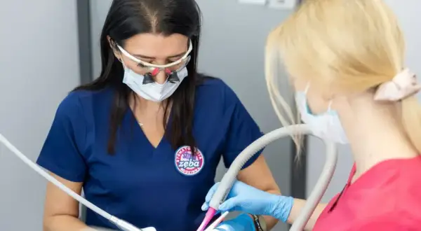 Dwie dentystki w maseczkach i okularach powiększających pracują przy pacjencie. Jedna używa wiertła, druga asystuje, podając anestetyki.