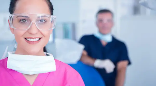 Uśmiechnięta asystentka dentysty w okularach ochronnych i różowym fartuchu, z maską na szyi. W tle widać lekarza.