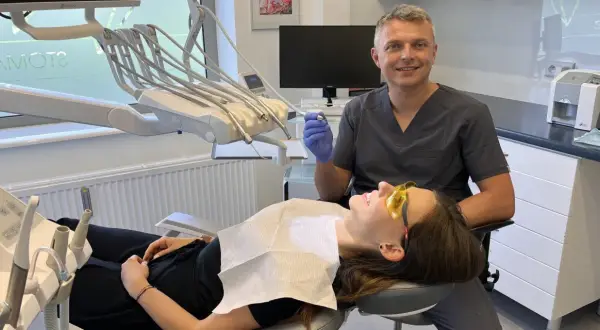 Dentysta w rękawiczce trzyma wiertło, pacjentka w okularach ochronnych leży na fotelu.
