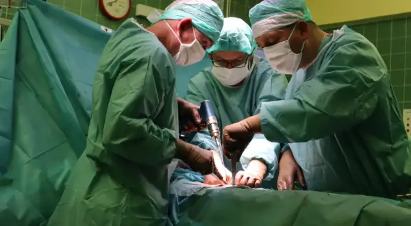 Trzech chirurgów w zielonych fartuchach i maseczkach wykonuje operację. Jeden z nich używa wiertarki chirurgicznej.