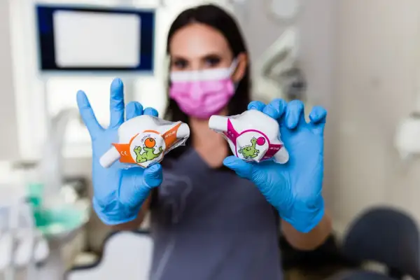 Dentystka w niebieskich rękawiczkach prezentuje dwa opakowania z zabawnymi dinozaurami, sugerujące zastosowanie w leczeniu, być może z użyciem podtlenku azotu.