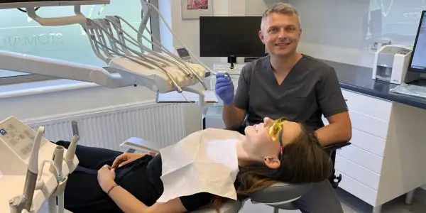 Dentysta w rękawiczce trzyma wiertło, pacjentka w okularach ochronnych leży na fotelu.